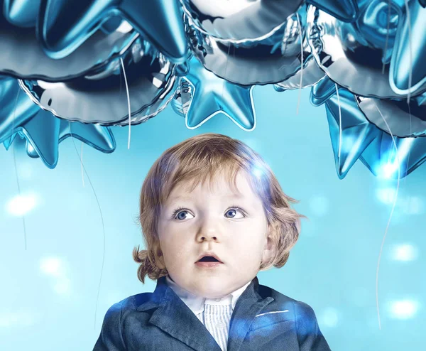 水色の背景の上に立っての青い風船を見てスーツでブロンドの髪と愛らしいびっくりした少年 — ストック写真
