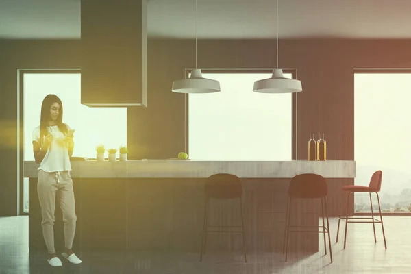 Woman in interior of gray kitchen with concrete floor, loft windows with moutain view and white and gray island with built in cooker and black stools. Toned image double exposure