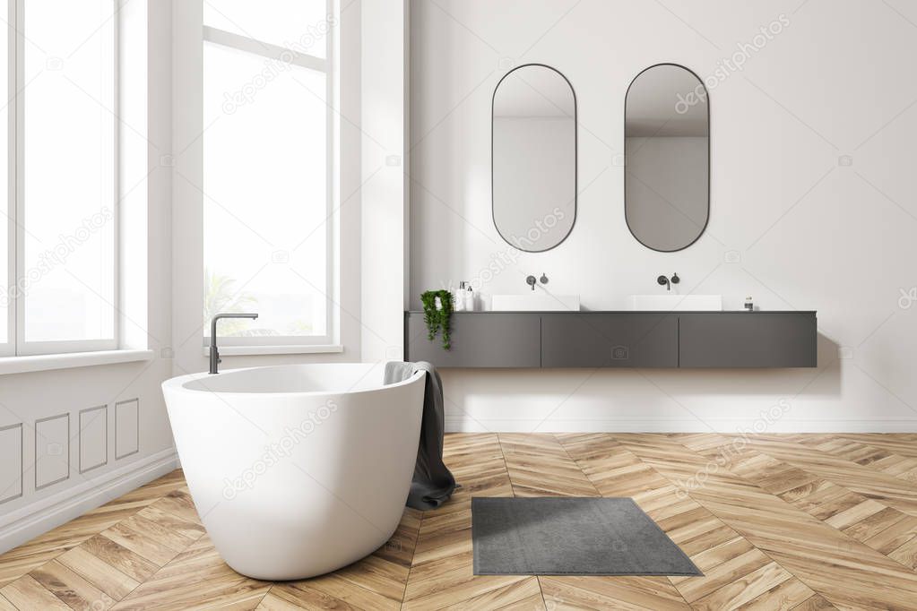 Interior of modern bathroom with white walls, wooden floor, white bathtub with a towel on it and two white sinks on gray counter with oblong mirrors above them. 3d rendering