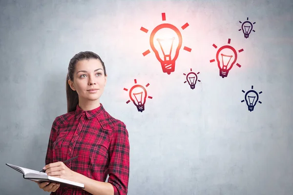 Young woman in checkered shirt holding copybook and pen standing near concrete wall with light bulbs drawn on it. Concept of idea