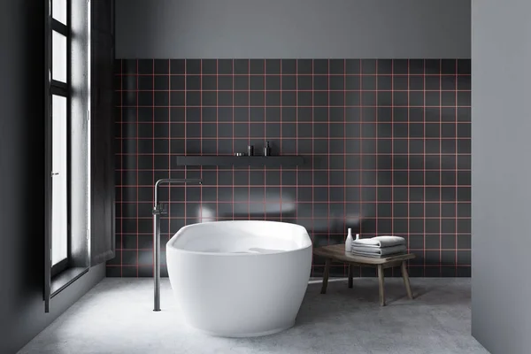 Interior of minimalistic bathroom with gray and black tile walls, concrete floor, white bathtub standing near window and black shelf with creams. 3d rendering