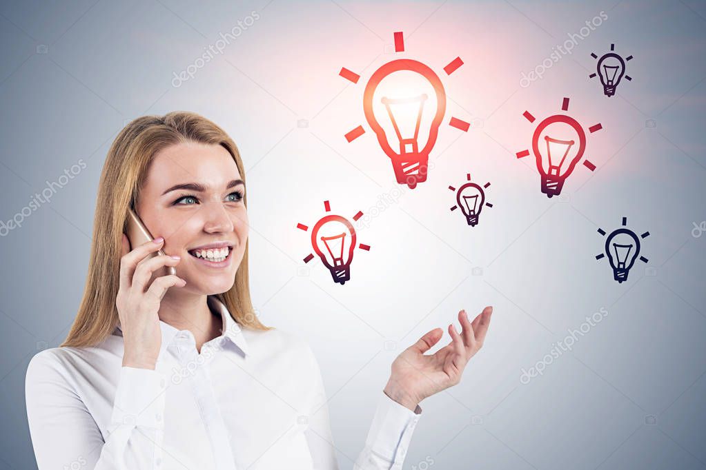 Smiling blonde businesswoman talking on smartphone standing near gray wall with light bulbs drawn on it. Concept of business idea