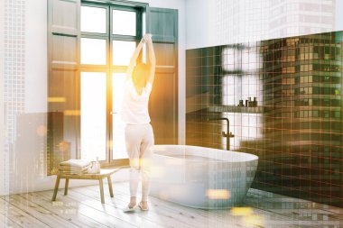 Kadın beyaz ve siyah taş duvarlar, ahşap zemin, pencere ve kremler ile siyah raf yakınındaki ayakta beyaz küvet ile banyo köşesinde. Çift tonlu görüntü pozlaması