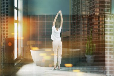 Gri ve siyah taş duvarlar, ahşap zemin ve pencerenin yakınındaki büyük gri küvet banyo içinde duran kadın. Çift tonlu görüntü pozlaması