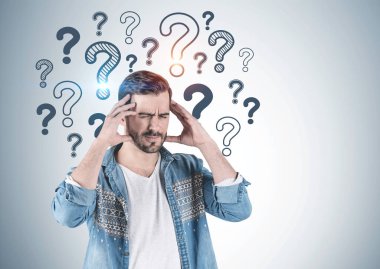 Soru işareti üzerinde çizilmiş olan kot gömlek ve sahip güçlü baş ağrısı ayakta gri duvarının yakınında giyen sakallı genç adam portresi. Yukarı alay