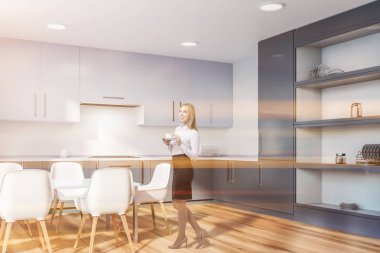 Kadın beyaz duvarlar, ahşap zemin, koyu gri mutfak tezgahı ve dolapları ile şık mutfak ve beyaz masa sandalye ile köşesinde. Tonlu görüntü