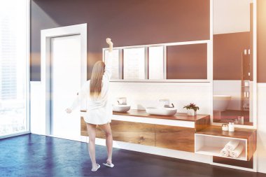 Kadın beyaz ve gri duvarlar, beton zemin, çift lavabo ayakta üstündeki dikey ve yatay ayna ile ahşap tezgah üzerinde ve bir kapı ile modern banyo köşesinde. Tonlu görüntü