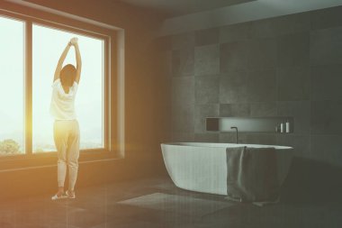 Kadın onu ve gri halı gri ve siyah taş duvarları, geniş pencere, gri havlu ile beyaz küvet ile şık banyo köşesinde. Çift tonlu görüntü pozlaması