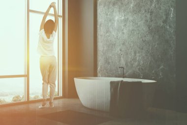 Kadın onu ve yakınında gri halı gri ve beton duvarlar, beton zemin, geniş pencere ve havlu ile beyaz küvet ile minimalist banyo köşesinde. Çift tonlu görüntü pozlaması