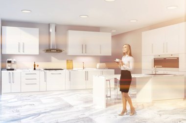 Kadın bej duvarlı modern mutfak köşesinde beyaz mutfak tezgahı ile yerleşik lavabo ve fırın, dışkı ile beyaz çubuğu ve dolaplar iki fırın içinde inşa ile. Tonlu görüntü