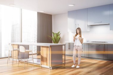 Kadın beyaz duvarlar, ahşap zemin, gri mutfak tezgahı ve dolapları ile panoramik mutfak köşe ve beyaz ve ahşap bar tabureleri ile. Tonlu görüntü