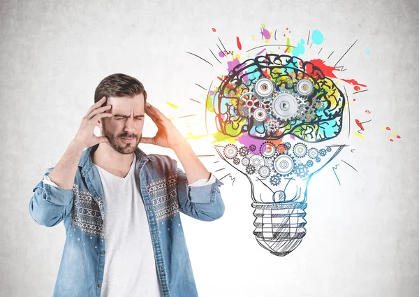 Side view of bearded young man in jeans shirt thinking hard with his eyes closed standing near concrete wall with cog brain and lightbulb drawn on it.