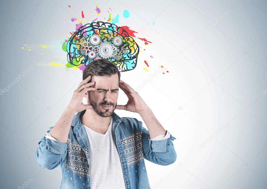 Portrait of bearded young man wearing jeans shirt and having strong headache standing near gray wall with cog brain drawn on it. Mock up