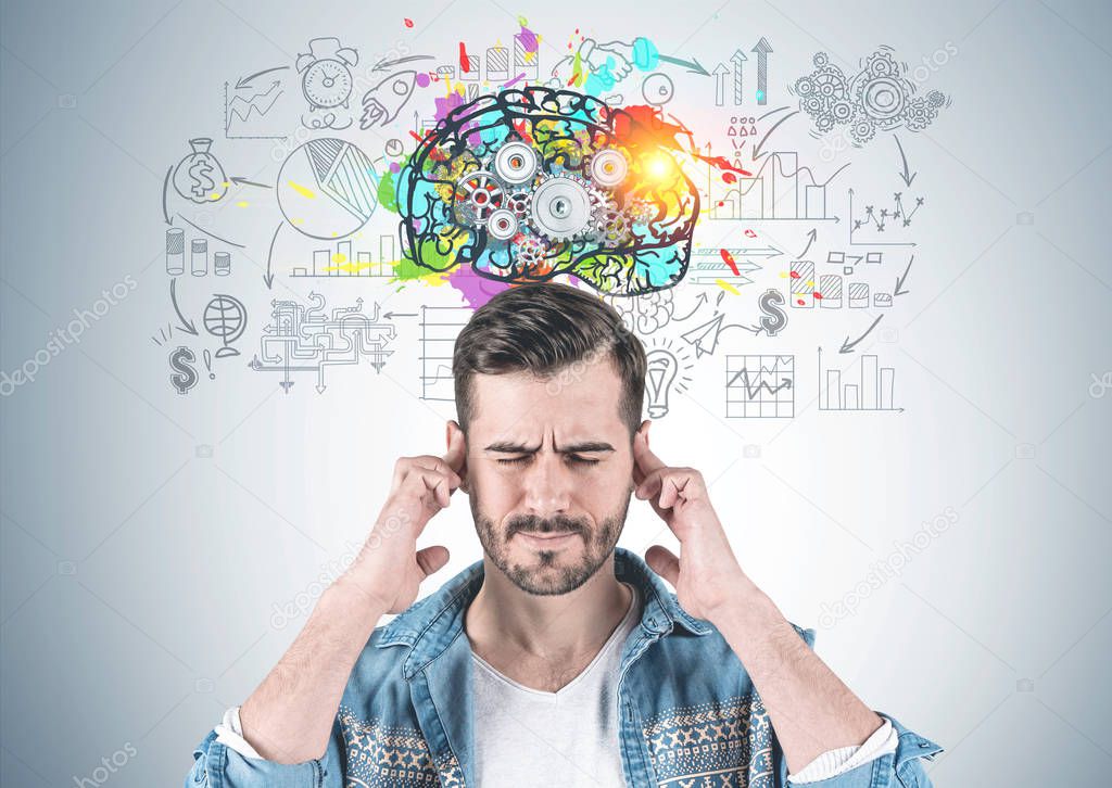 Portrait of bearded young man wearing jeans shirt and thinking hard with eyes closed and fingers on temples standing near gray wall with colorful start up sketch