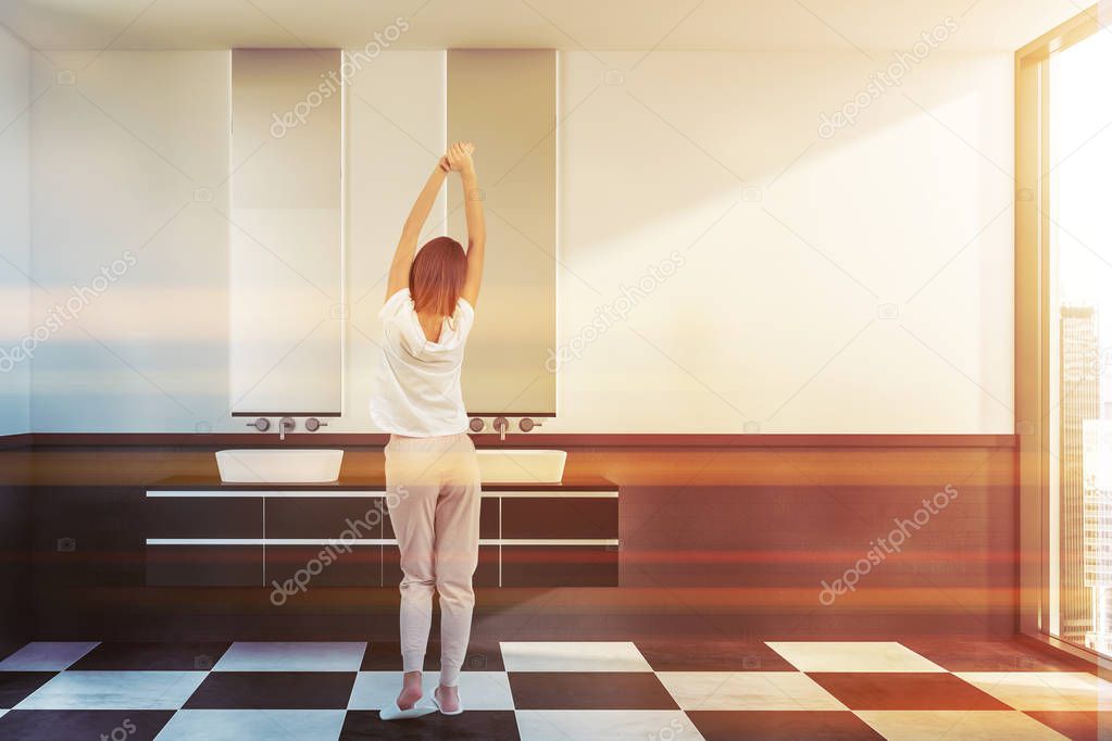 Woman in interior of stylish bathroom with white and gray brick walls, tiled floor, double sink standing on black and white countertop and two vertical mirrors. Toned image