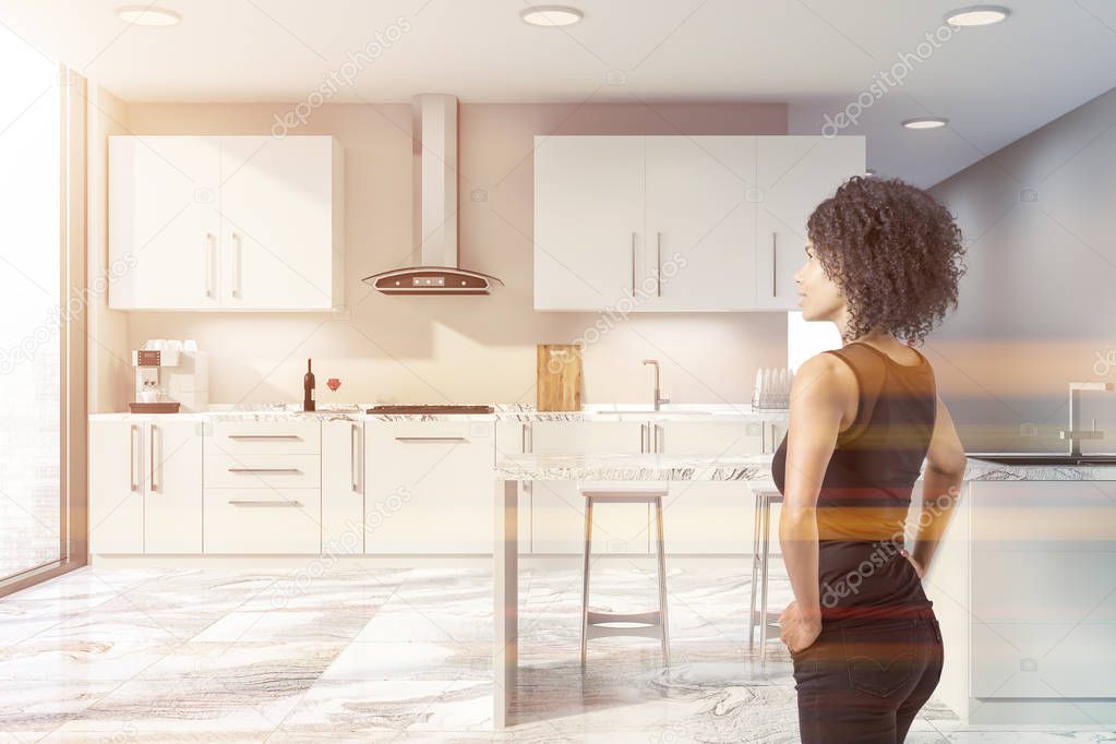 Woman in interior of stylish kitchen with beige walls, white marble floor, large windows, white countertops with built in sink and oven, coffee machine and white bar with stools. Toned image