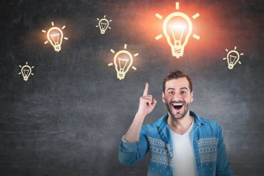 Kot gömlek ayakta açık ağız ve çevre yazı tahtası üzerinde çizilmiş sarı ışık ampuller ile işaret parmağı ile mutlu genç adam portresi. İyi fikir kavramı