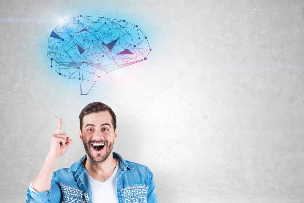 Portrait of happy young man in jeans shirt standing with open mouth and finger pointing up near concrete wall with polygonal brain sketch drawn on it. Concept of brainstorming. Mock up