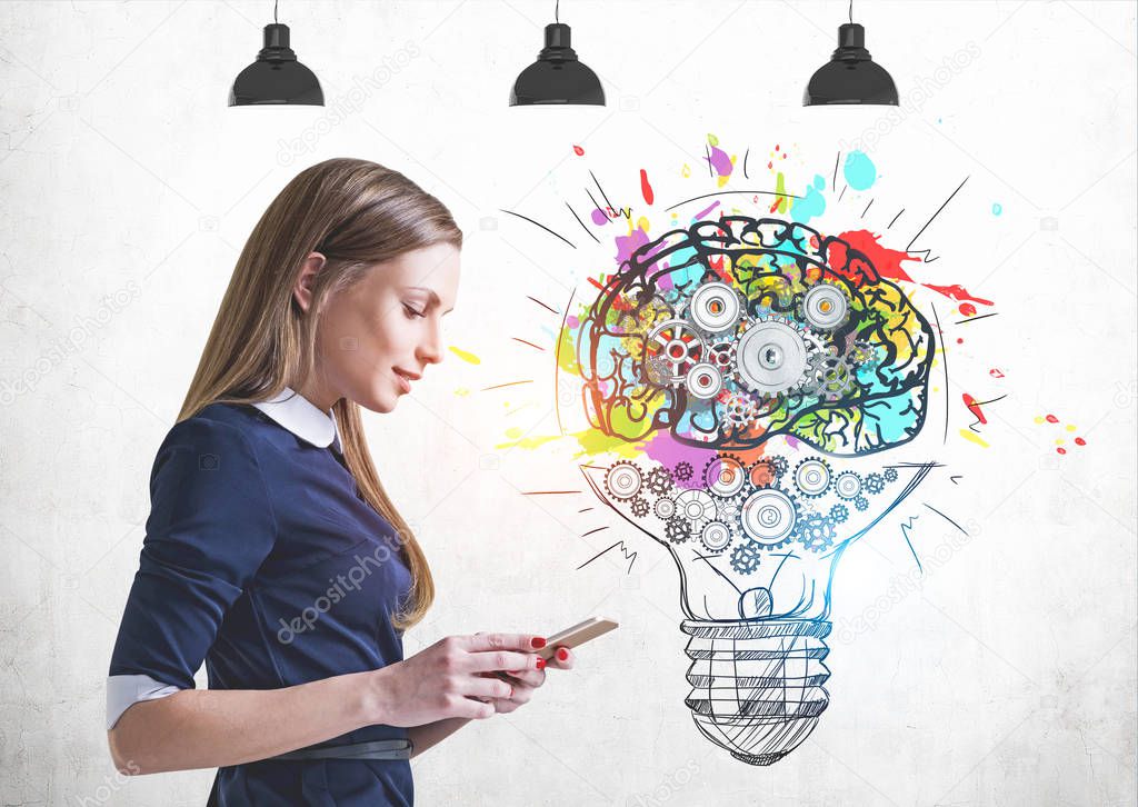 Beautiful blonde businesswoman looking at her smartphone standing near concrete wall with with cog brain and lightbulb drawn on it. Concept of creative thinking
