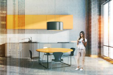 Kadın iç beton ve gri duvarlar, beton zemin, panoramik pencere, turuncu dolabı, modern mutfak, mutfak tezgahı ve yuvarlak masa sandalye ile gri. Çift tonlu görüntü pozlaması