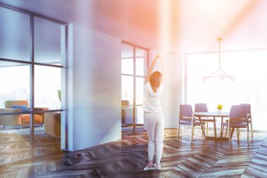 Kadın minimalist yemek odası ile beyaz duvarlar, ahşap zemin, geniş pencere, gri yuvarlak masa gri sandalyeler ve oturma odası sol köşesinde. Tonlu görüntü