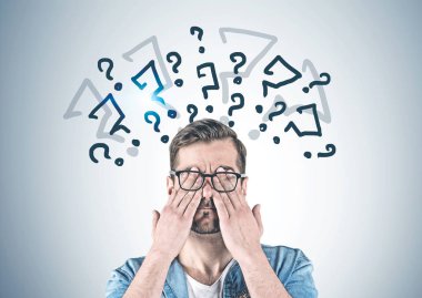 Rahat kıyafetler ve soru işareti üzerinde çizilmiş olan gri duvara yakın duran elleriyle gözlerini kapsayan gözlük sakallı genç adam portresi.