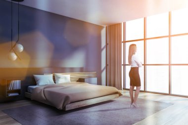 Sarışın genç bir kadın panoramik yatak odası köşesinde duruyor. Gri duvarları, ahşap zemini, rahat büyük boy yatağı, kitaplığı ve bulanık şehir manzarası var. Tonu ayarlanmış resim