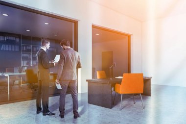 İki genç iş adamı, beyaz CEO ofisinde fayanslı zemini, turuncu sandalyeli kocaman masası ve arka planda açık alanı ile konuşuyor. Liderlik kavramı. Tonu ayarlanmış resim