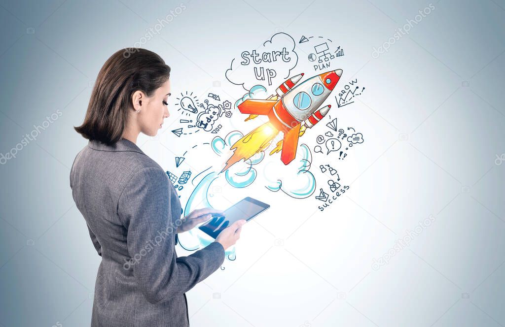 Side view of serious young European businesswoman using her tablet near gray wall with colorful startup sketch drawn on it. Concept of new project launch and technology