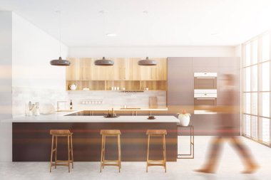 Beton zemini, gri tezgahları, tahta dolapları ve tabureli barları olan beyaz panoramik mutfakta yürüyen bulanık genç adam. Tonu ayarlanmış resim