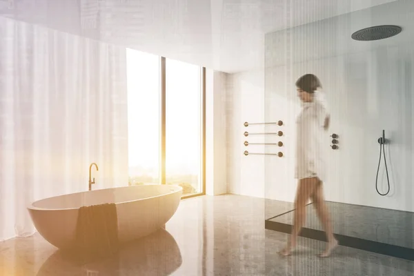 Blurry young woman in nightgown walking in luxury bathroom with white walls, concrete floor, comfortable bathtub and shower stall. Toned image double exposure