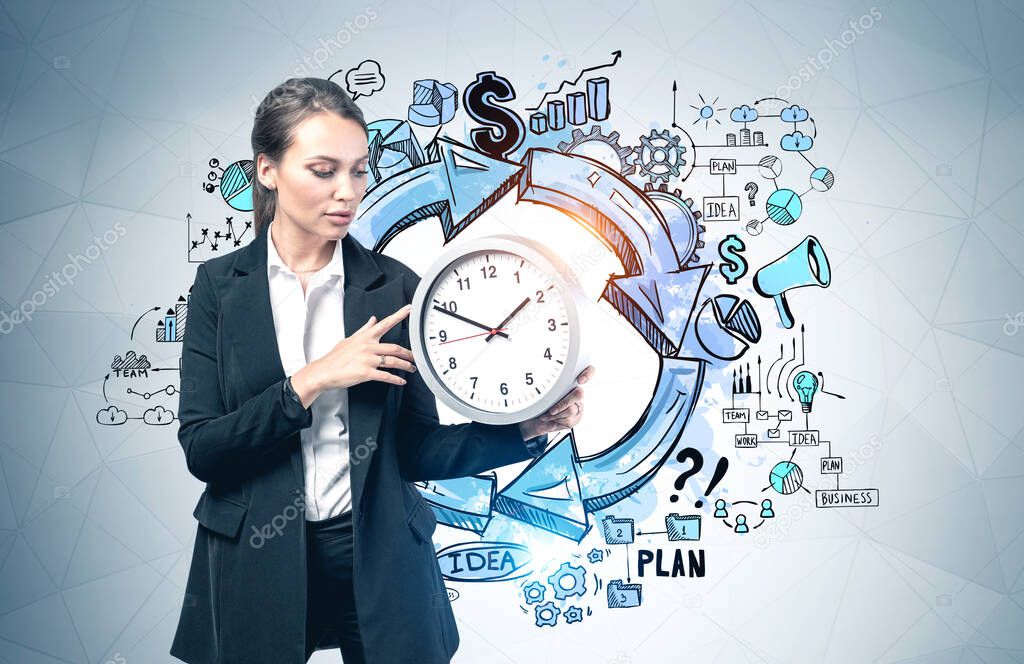 Serious young European businesswoman holding office clock near gray wall with creative business sketch drawn on it. Concept of planning and time management
