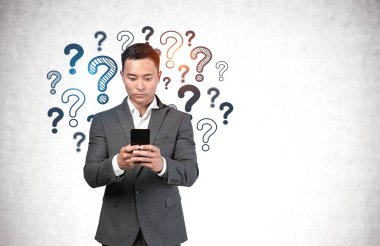 Ciddi genç Asyalı işadamı beton duvarın yanında akıllı telefon kullanıyor ve üzerinde soru işaretleri var. Seçim ve internet arama kavramı. Model