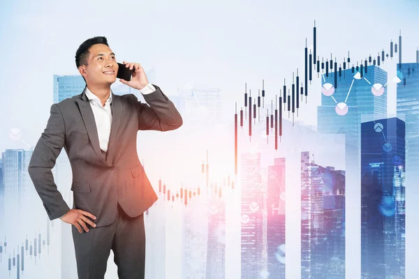 Portrait of smiling young Asian trader talking on smartphone in blurry abstract city with double exposure of financial graph. Concept of stock market. Toned image