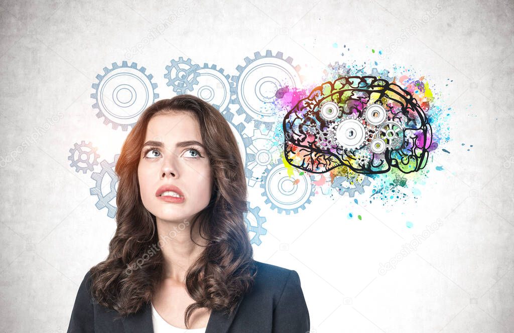 Portrait of troubled young businesswoman with wavy dark hair standing near concrete wall with colorful brain sketch drawn on it. Concept of brainstorming and problem solving