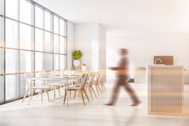 Takım elbiseli bulanık genç adam geniş panoramik yemek odasında beyaz duvarlı, beyaz sandalyeli uzun bir masa ve arka planda televizyon ve kanepe olan oturma odasında yürüyor. Tonu ayarlanmış resim