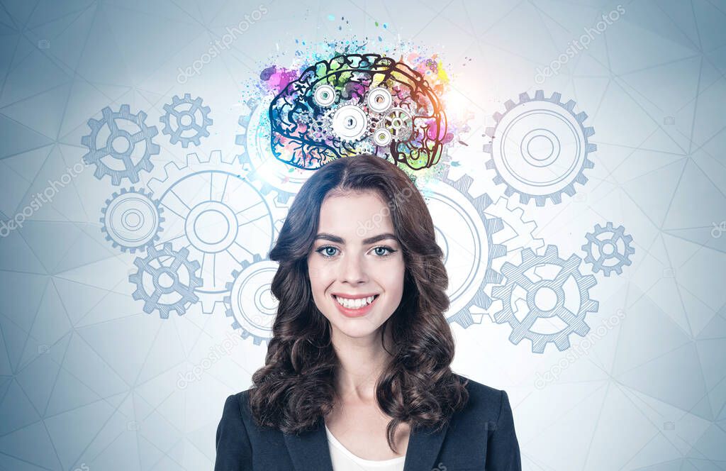 Happy young European businesswoman with wavy dark hair standing near gray wall with colorful brain with gears drawn on it. Concept of brainstorming and education