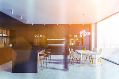 Geniş bir mutfakta siyah ve ahşap duvarları, tabureleri ve beyaz yemek masası olan bar tezgahı ve şehir manzaralı balkonu olan bulanık genç bir kadın. Tonu ayarlanmış resim