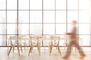 Takım elbiseli bulanık genç adam panoramik yemek odasında ahşap zemin ve beyaz sandalyeli uzun bir masa ile yürüyor. Modern şehir manzaralı bir pencere. Tonu ayarlanmış resim