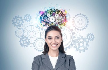 Üzerinde vitesler çizilmiş renkli beyin çizimi olan gri duvarın yanında duran koyu saçlı neşeli Avrupalı genç iş kadını. Beyin fırtınası kavramı