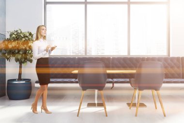 Beyaz duvarları, ahşap zemini, mavi koltuğu, sandalyeleri ve ahşap masası olan modern restoranda kahve içen sarışın kadın. Bulanık şehir manzarası. Tonu ayarlanmış resim