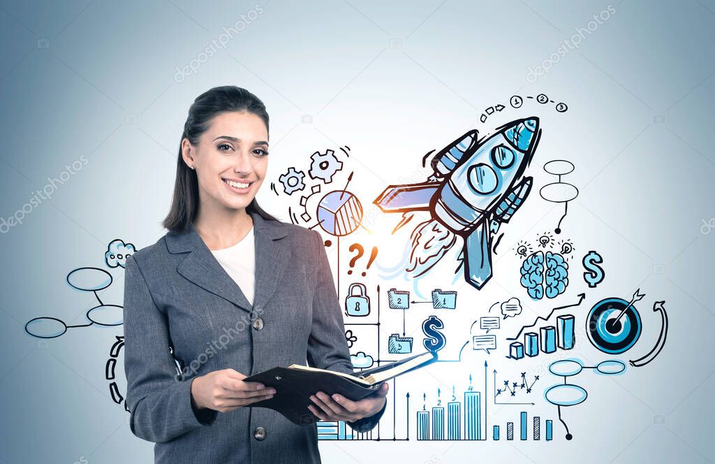 Cheerful young European businesswoman with dark hair holding notebook near gray wall with creative startup sketch. Concept of business planning, innovation and new project launch