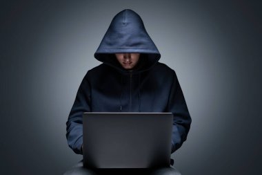 Siyah kapüşonlu genç hacker 'ın portresi gri duvar arkasında dizüstü bilgisayarını kullanıyor. Siber güvenlik ve veri koruması kavramı
