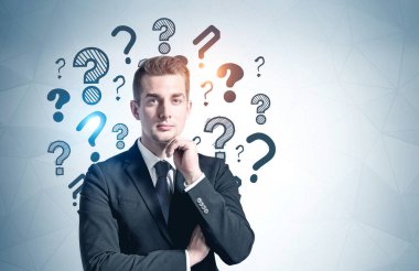 Üzerinde soru işaretleri olan gri duvarın yanında duran düşünceli genç işadamı. Model