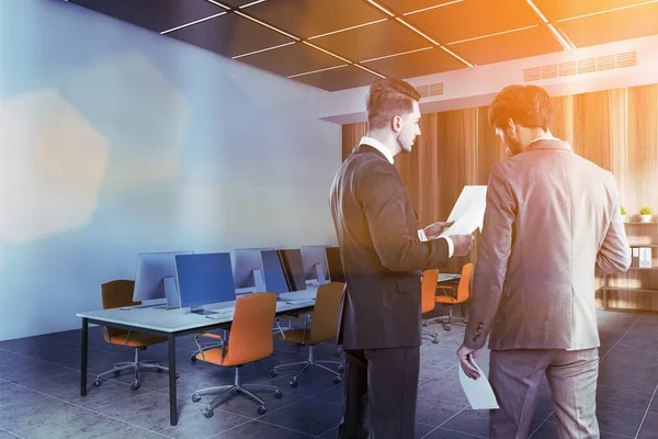 Two businessmen discussing documents in open space office with white and wooden walls and long computer table. Toned image