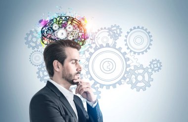 Üzerinde çarklar çizilmiş gri duvarın yanında duran takım elbiseli dalgın sakallı genç adamın yan görüntüsü. Beyin fırtınası kavramı