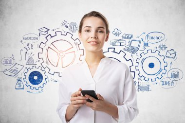 Akıllı telefonlu gülümseyen Avrupalı genç iş kadını yaratıcı vites çizimiyle beton duvarın yanında duruyor. Takım çalışması kavramı.