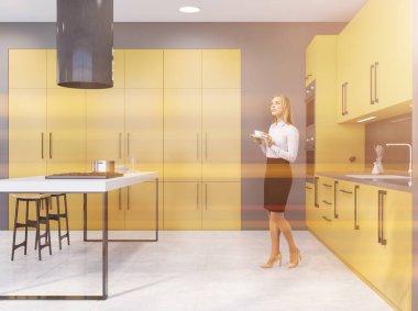 Gri duvarları, sarı dolapları ve tabureli barları olan modern mutfakta duran güzel genç bir kadın. Tonu ayarlanmış resim