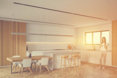 Mutfak köşesinde beyaz ve ahşap duvarları, beyaz dolapları, bar ve yemek masası olan genç bir kadın. Renkli resim çift pozlama