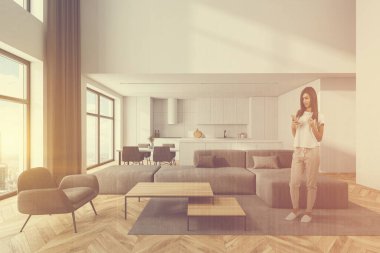 Beyaz duvarları, ahşap zemini, gri koltuğu, arka planda koltuk ve mutfağı olan modern oturma odasında duran genç bir kadın. Renkli resim çift pozlama